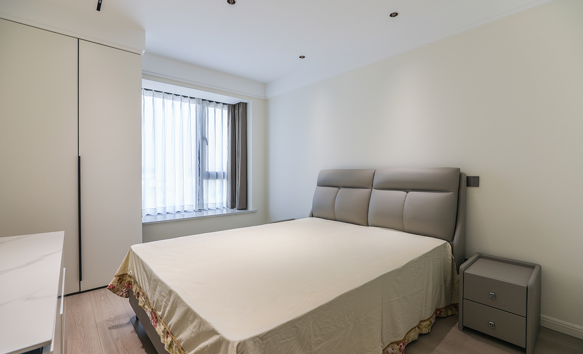 次臥地面同樣鋪以暖灰色地板，與其他房間相呼應(yīng)，窗簾的色彩與床的色彩接近，體現(xiàn)了主人在軟裝上的極致追求。衣柜靠窗，不落俗套，讓空間布局更合理適宜，白色臺面床頭柜，整潔大方，配上吊頂里鑲嵌的筒燈，讓人有一種心曠神怡的感覺。