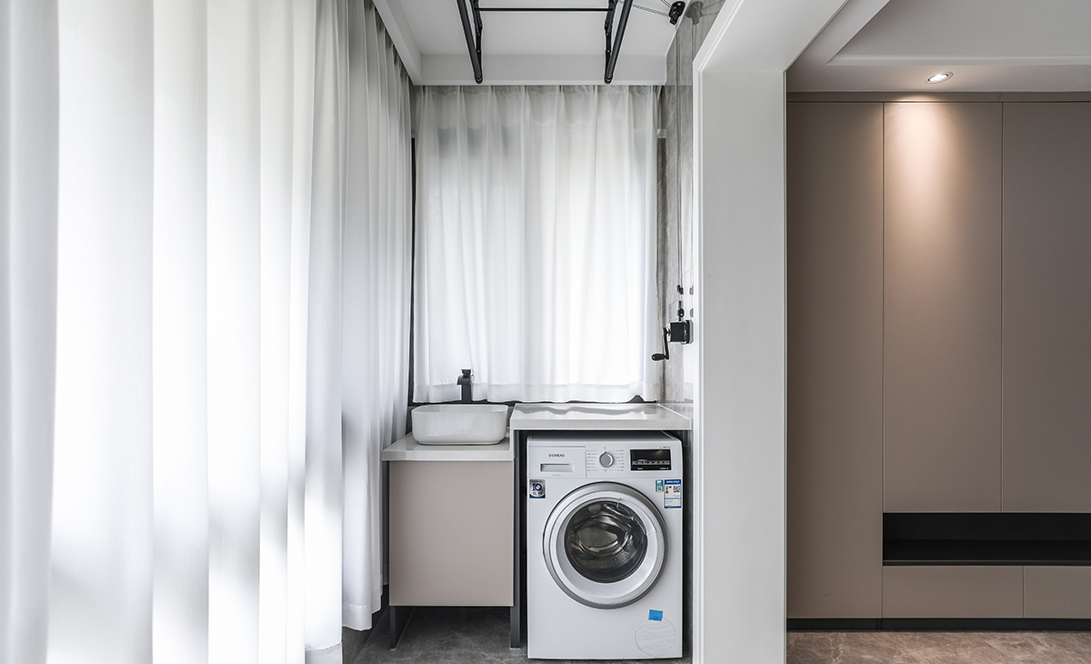 陽臺落地玻璃，洗衣柜的一面砌了矮墻，方便排水管，整體更美觀。陽臺的承重墻兩邊對應著一面儲物柜，一面洗衣機臺盆組合柜，簡約大氣，功能合理分配。