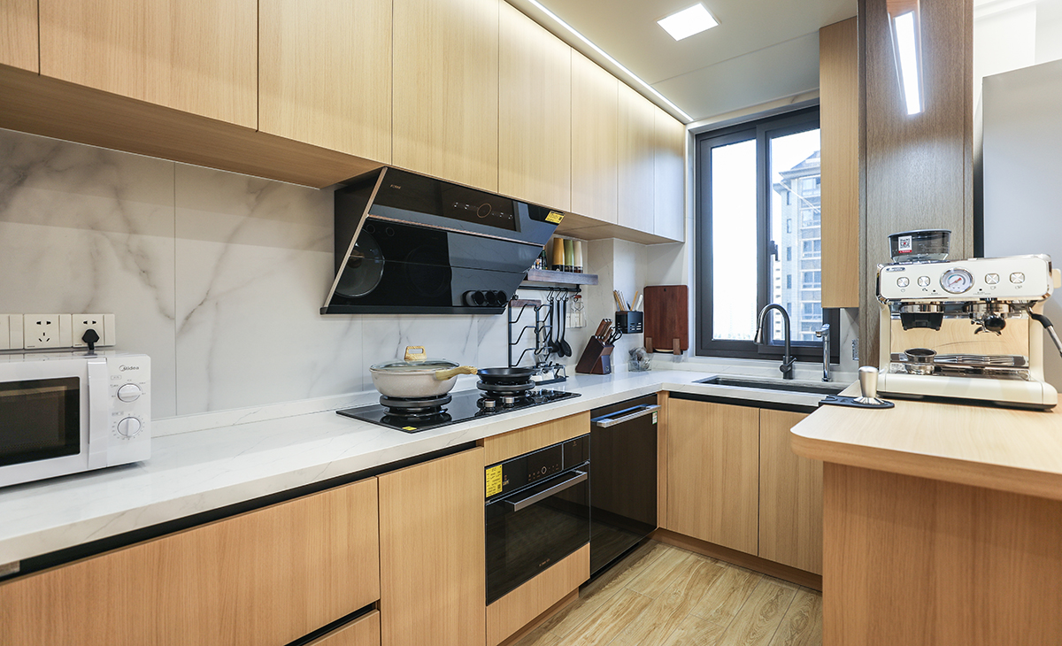 廚房間本來是長方形封閉式的空間，改成開放式的廚房，配上吧臺，L型櫥柜整體采用原木色門板，配上白色的臺面，整體干凈、簡潔，地面用的木紋磚，與廚柜門板和吧臺顏色統一，頂面采用現在流行的大板吊頂配上線性燈，時尚、溫馨。