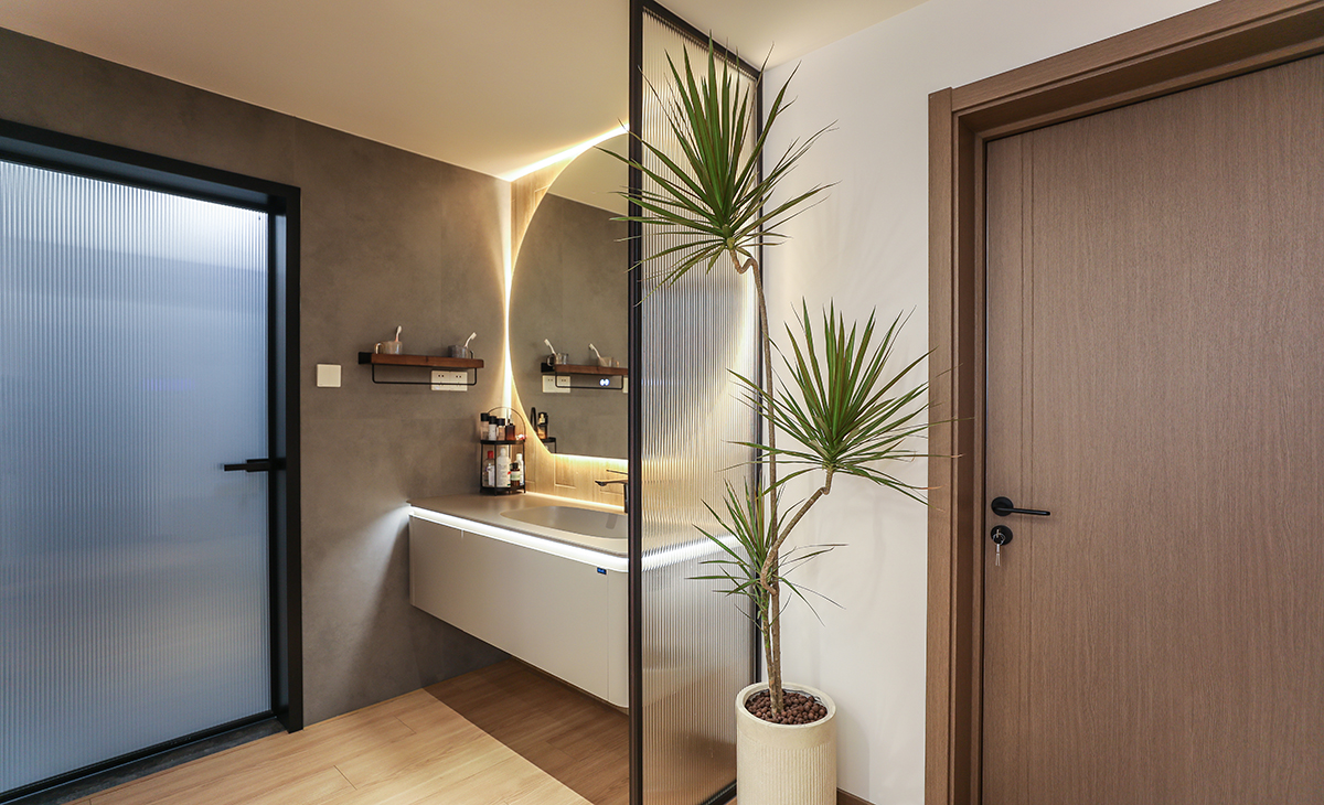 進門口中間正好是個走道，做了個干濕分離，用現在流行的長虹玻璃做隔斷，透光而有私密感，墻面采用黑色水泥磚，用原木色木紋磚上墻，氛圍感一下就拉滿了，圓形的鏡子配上線性燈，整體效果圖凸出，時尚、大氣、簡單。