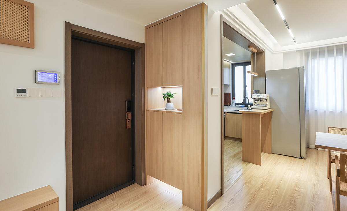 進門口地面鋪的實木復合地板，左手邊做了原木色鞋凳，上面的配電箱也選擇了原木色的裝飾畫作為隱藏，右手邊做了一個收納鞋柜，中間和下面做鏤空，原木色的柜體和門板配上線性燈，更簡潔、明亮。