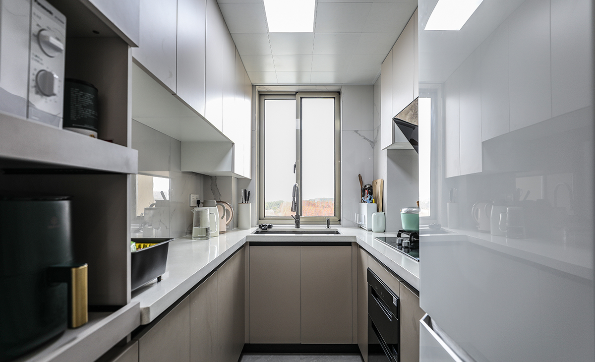 該套房子廚房設計U型櫥柜，增加操作臺面，一邊40cm深淺柜，用來放各種設備，顏色搭配上淺下深，石英石材質(zhì)臺面更好打理，空間劃分合理，滿足主人制作各種美食的需求，原櫥門拆除，做三聯(lián)玻璃移門，整體進出更加方便，空間顯明亮。