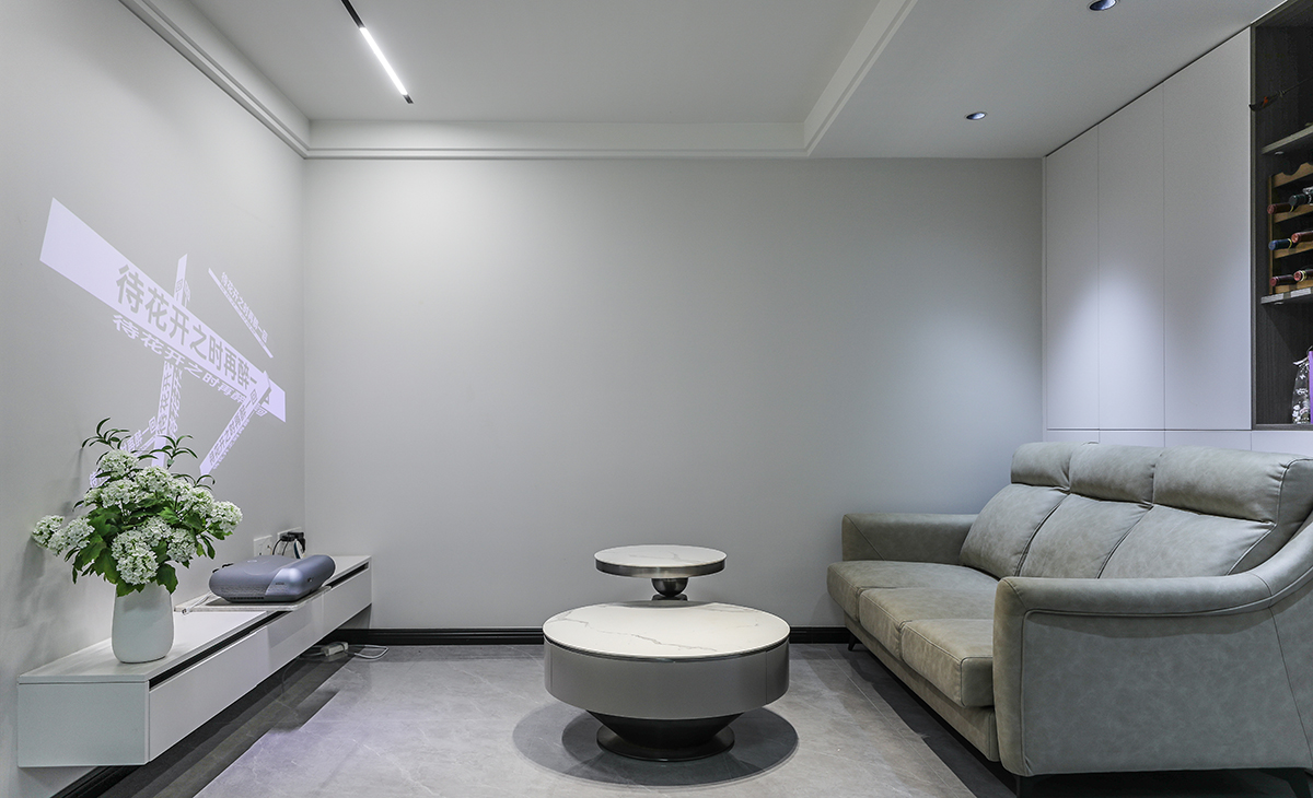  客廳開間很大，電視墻做白色乳膠漆墻面用做投影幕布?jí)Γ浜蠏靿κ诫娨暪瘢嘲l(fā)背景白色門板配深灰開放隔顏色相互配合，明暗有神，淺灰色皮質(zhì)沙發(fā)立刻檔次提高很多，放一組高低巖板茶幾柜，客廳彰顯靈動(dòng)。吊頂造型上采用雙眼皮無主燈吊頂。地面采用淺灰色地磚。造型和色調(diào)上無不給人以簡(jiǎn)潔、大氣、時(shí)尚的感覺。