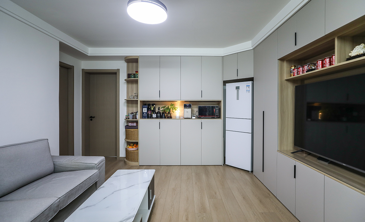  客廳開間很大，電視柜于展示柜淺色的門板和原木色木飾面相互配合，明暗有神，深灰色皮質沙發立刻檔次提高很多，客廳空間也很寬闊。吊頂造型上采用雙眼皮吊頂。地面原木色地板。造型和色調上無不給人以簡潔、舒適、溫馨的感覺。