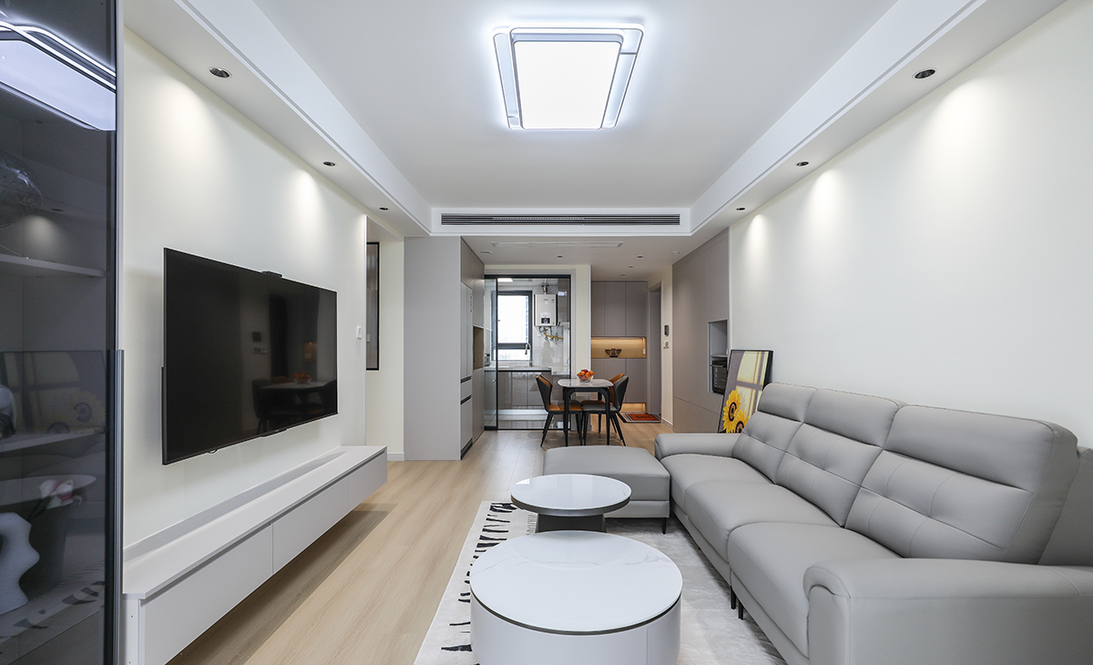  客廳面積不大，但經過設計師的手筆，讓空間緊湊不擁擠，電視墻做展示立柜與懸空電視柜組合，柜子色系和沙發相互配合，明暗有神，深灰色皮質沙發立刻檔次提高很多。吊頂造型上采用雙眼皮圍邊吊頂，讓造型簡介配合筒燈打造氛圍感。地面采用原木色地板。造型和色調上無不給人以簡潔、溫馨、時尚的感覺。