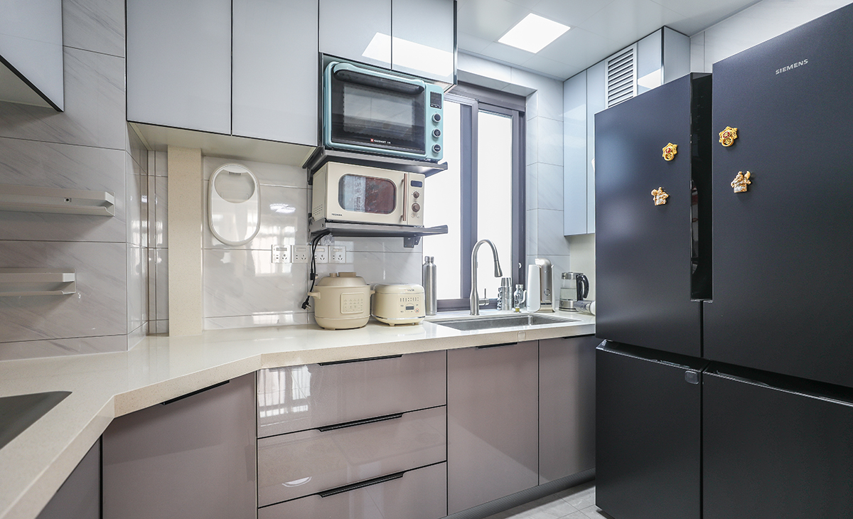 該套房子廚房空間很大，L型三面櫥柜，另外冰箱邊上也有操作臺(tái)，顏色簡(jiǎn)潔明快，材質(zhì)要干凈和好打理，白色的吊頂和米色的上櫥柜、深色的下櫥柜相呼應(yīng)，石英石臺(tái)面干凈、好打理，各種廚房電器全部配齊，滿足主人制作各種美食的需求。