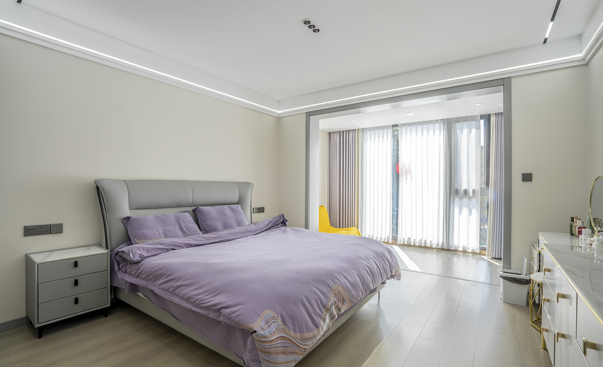  主臥空間很大，頂面有主光源，有輔助光線和氛圍燈，把空間烘托的很浪漫、很溫馨，原木色地板搭配可可蛋奶墻面涂料，灰色的門套和灰色的床相呼應，加上紫