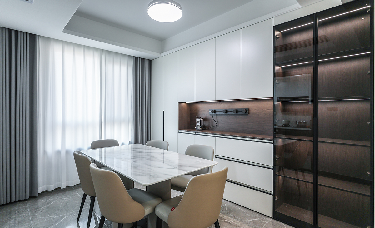 餐廳空間比較方正寬敞，又有一個大飄窗，靠墻做一排裝飾柜，既實用、又有裝飾功能，原木色框架，極窄邊框玻璃門和白色木門板相呼應，巖板餐桌把用餐氛圍調和的很溫暖，淺灰色地磚很干凈，色彩和造型上都簡潔大氣。