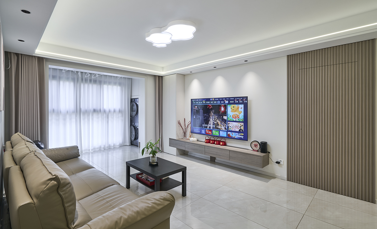 客廳開間很大，電視墻用木格柵作為主臥隱形門，I型的懸空電視柜，咖色皮質沙發立刻檔次提高很多，放一組三人妃沙發，客廳空間也很寬闊。頂面中央空調，吊頂造型上采用雙眼皮吊頂。地面采用暖灰色地磚。造型和色調上無不給人以簡潔、大氣、時尚的感覺。