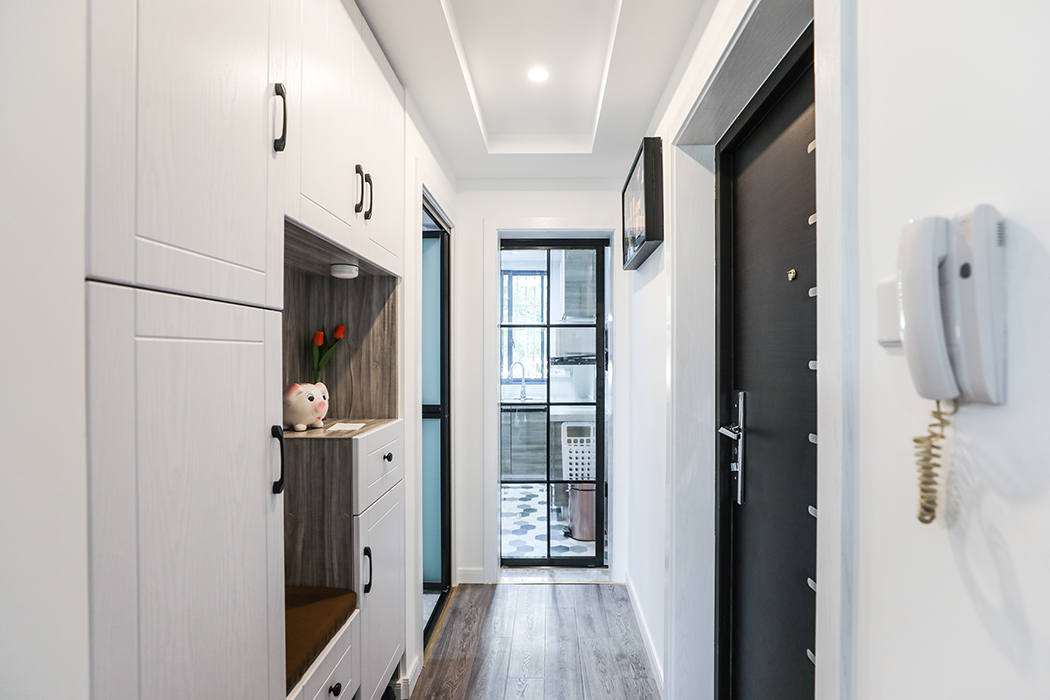 將鞋柜與其他收納空間整合至一側(cè)墻面，視線前方廚房門運用簡約幾何形元素，光線自然穿過狹小過道，使小屋充滿溫馨感。
