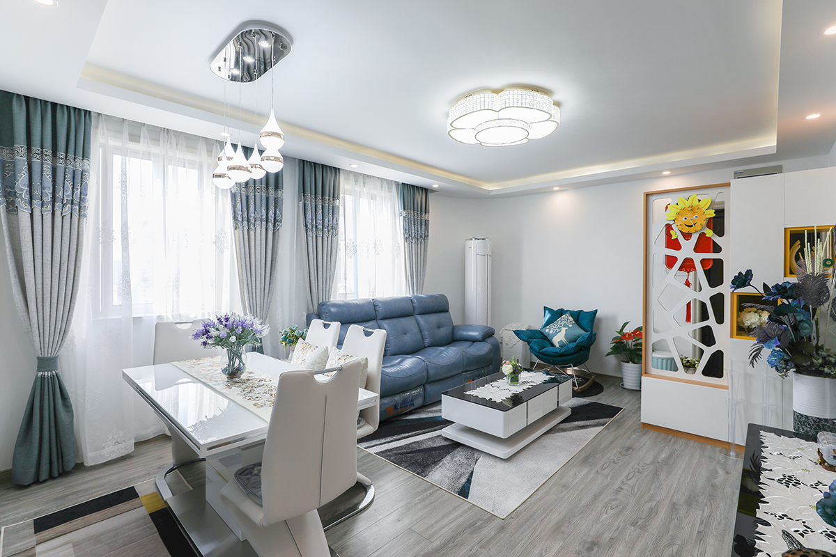 客餐廳頂面平頂，四周做造型頂，配以燈槽及筒燈作為氛圍燈。墻頂面白色乳膠漆干凈簡潔。地面鋪裝深色地板，色彩重心平穩(wěn)。淺藍(lán)色窗簾及藍(lán)色沙發(fā)相近色配色顯得空間既清爽又有層次感。
