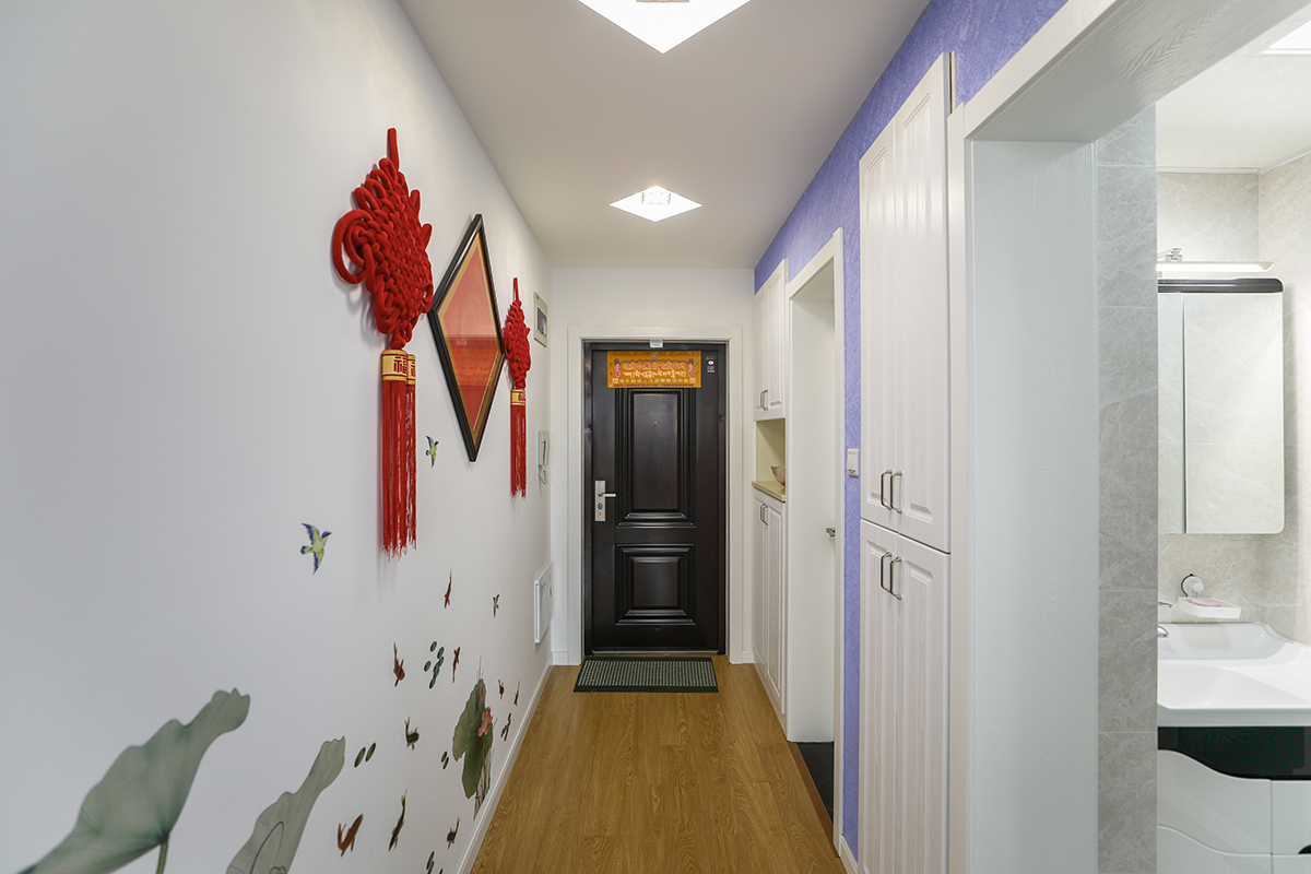 入門的玄關沒有過多的修飾，大門的門套結合右邊的鞋柜做了加寬的處理，這樣在視覺上更為整體，不會讓入門這塊形成轉折的感覺，內門套是不銹鋼包邊，另一面的墻壁上掛著喜慶中國結，下方水墨裝飾畫使公共開放區域成為整體貫穿元素的有機聯系。