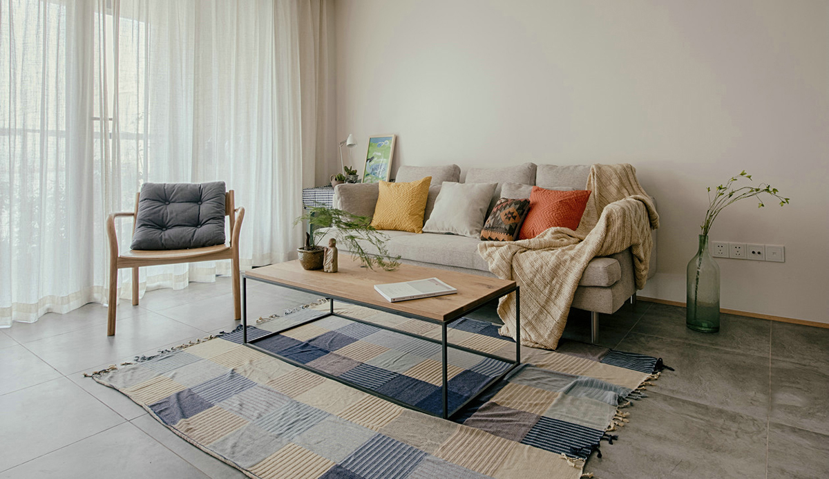 客廳空間開闊明亮。利用淺色紗幔窗簾優化自然光的吸采，讓空間擁有足夠的恬靜感。業主對日式風格很喜歡，客廳的茶幾是業主在日本旅游時帶回國。灰色的布藝沙發，沉穩大氣，搭配自然紋理的原木茶幾和座椅，讓濃郁的日式風情填滿整個空間。柔軟舒適的沙發，隨意擺放著幾個抱枕，再增加一張橙色的真皮單人椅，無形之中提升了空間的質感。
