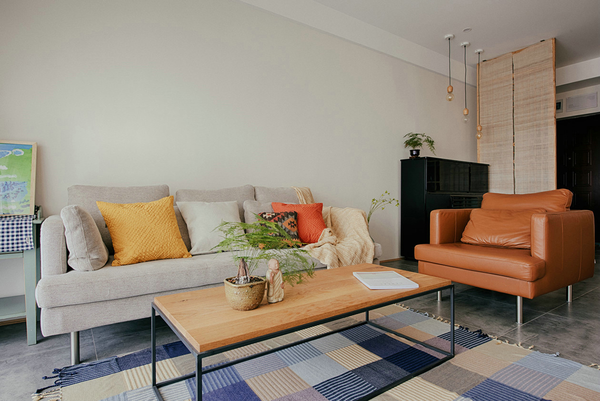 客廳空間開闊明亮。利用淺色紗幔窗簾優化自然光的吸采，讓空間擁有足夠的恬靜感。業主對日式風格很喜歡，客廳的茶幾是業主在日本旅游時帶回國。灰色的布藝沙發，沉穩大氣，搭配自然紋理的原木茶幾和座椅，讓濃郁的日式風情填滿整個空間。柔軟舒適的沙發，隨意擺放著幾個抱枕，再增加一張橙色的真皮單人椅，無形之中提升了空間的質感。
